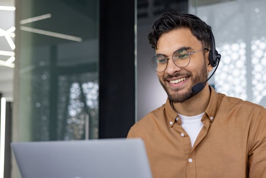 Smiling customer service representative wearing a headset and glasses, working on a laptop in a modern office space. photo