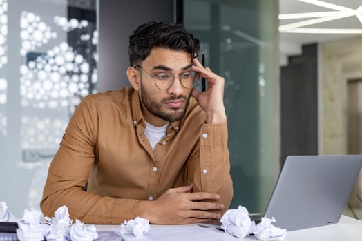 Stressed businessman wearing glasses and brown shirt working at desk with crumpled papers and laptop in modern office environment. photo