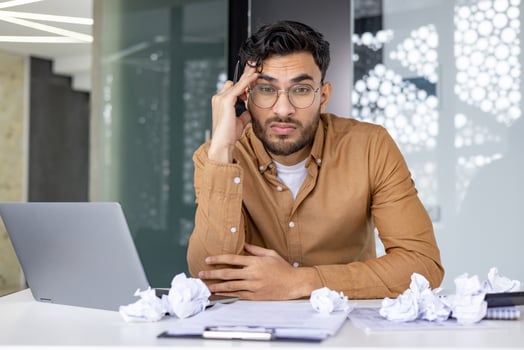 Young businessman looking frustrated at his desk with a laptop and crumpled papers, representing stress and deadlines in a modern office setting. photo