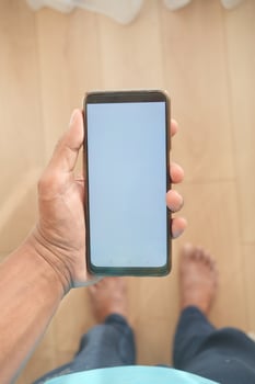 A hand is holding a smartphone with a blank screen on a wooden floor in a closeup shot photo