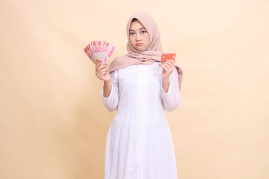 girl asian muslim in hijab frowning holding rupiah cash and credit card in hand. Lifestyle, religious, business and finance concepts photo
