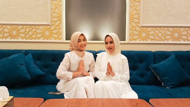 a pair of women sitting on a sofa smiling gracefully with a welcome hand gesture sawadikap facing the camera with an elegant Arabic Muslim calligraphy background photo