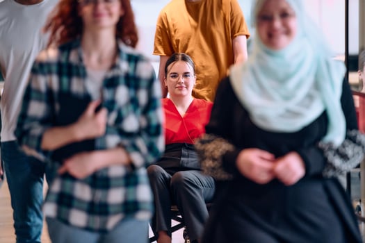 A diverse group of young business people walking a corridor in the glass-enclosed office of a modern startup, including a person in a wheelchair and a woman wearing a hijab photo