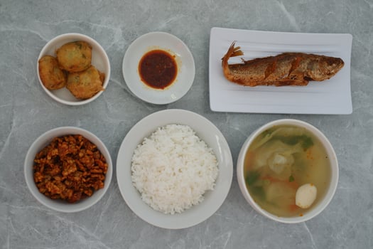 A meal consisting of a plate of fried fish, a bowl of white rice, a bowl of vegetable soup with meatballs, a small plate of red sauce and others, all on a gray marble surface. photo