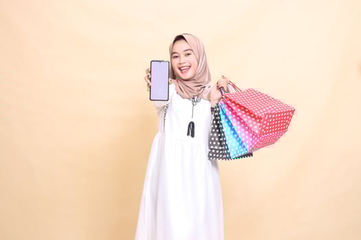 a young Asian Muslim woman wearing a hijab happily displays a cellphone screen and carries a paperbag shopping bag. for advertising, technology, Eid and Ramadan photo