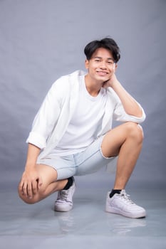 elegant smiling asian man posing in upright squats in front of the camera while holding his hands to his cheeks indoors. fashion promotion man shirt photo session in the gray background studio