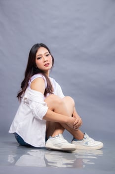 elegant woman posing sitting in front of the camera while her ladder holding her feet indoors. fashion promotion woman shirt photo session in gray background studio