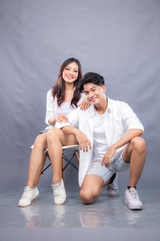 smiling man and woman posing sitting in front of the camera while their ladders holding their thighs facing forward indoors. fashion promotion couple photo session in gray background studio