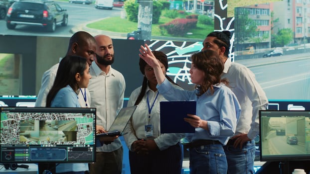 Diverse employees group discussing ways to speed up order delivery, looking for routes to avoid traffic on satellite navigation. Agency operators use CCTV footage in monitoring room. Camera A. photo