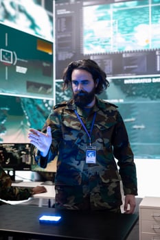 Army soldier examines real time data on a hologram 3d projection, reviewing defense operation details in military control room. Trooper looks at CCTV satellite footage on 3d visualization. photo