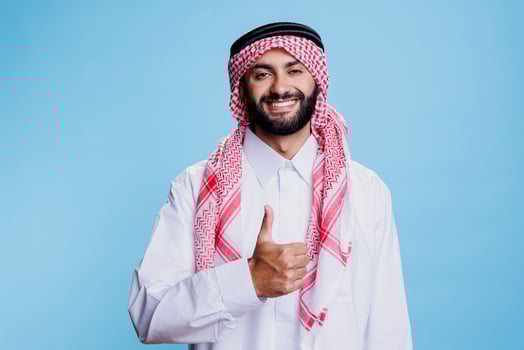 Smiling muslim man in white thobe and checkered headdress showing thumb up studio portrait. Cheerful arab posing with positivity and satisfaction gesture and looking at camera photo
