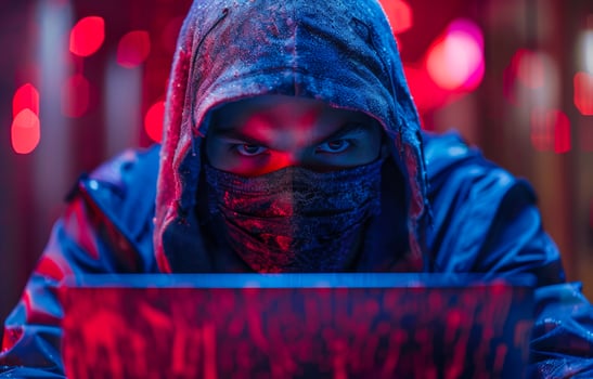 Masked Hacker Working On Laptop At Night. A hooded figure wearing a face mask and looking intently at a laptop screen. photo