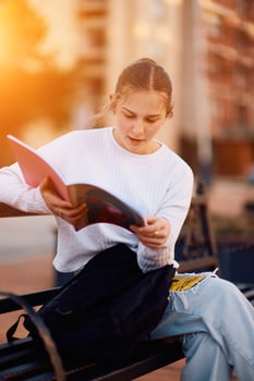 A beautiful blonde teenager sits on a bench in the park, taking a break from school to immerse herself in a book, accompanied by her school backpack photo