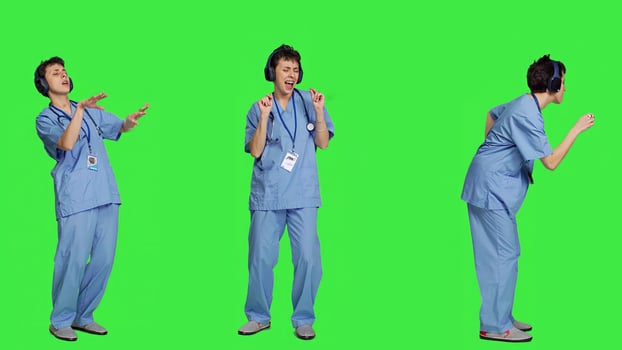 Nurse having fun listening to music on wireless headphones, wearing scrubs and dancing around against greenscreen backdrop. Medical assistant showing groovy funky dance moves. Camera A. photo