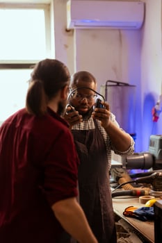 Master in carpentry shop instructing BIPOC worker to wear protection gear, preventing workplace accidents. Cabinetmaker making sure apprentice uses safety earmuffs and goggles to avoid injury photo