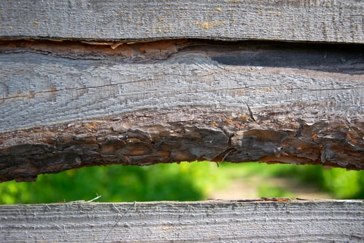 A close-up view of a weathered wooden fence with a small gap revealing a blurred green landscape photo