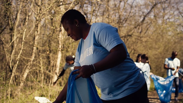 African american activist doing litter cleanup to fight illegal dumping, protecting the natural environment with garbage disposals. Woman working on collecting rubbish from the forest area. Camera B. photo