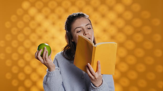 Woman reading book and enjoying fresh apple, living healthy, isolated over studio background. Bookworm entertained by novel eating green fruit, enjoying bio snack, camera B photo