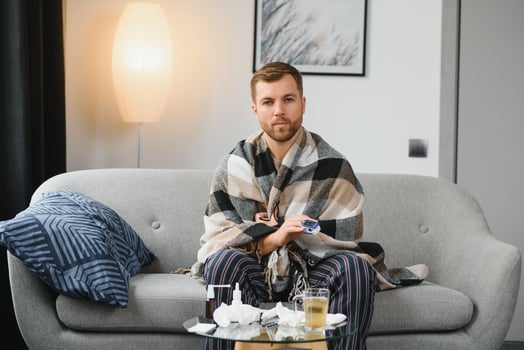 Sick adult man who has bad cold or seasonal flu virus is fighting illness at home. Unhappy middle aged man is sitting on sofa wrapped in warm scarf and plaid, holding paper tissue and sneezing. photo
