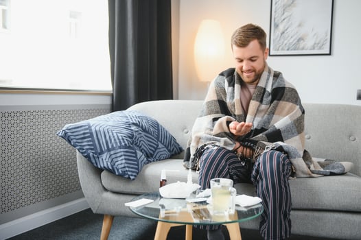Sick bearded man who has bad cold or seasonal flu sitting on couch at home. Guy with fever wearing warm plaid shivering with worried face expression. photo