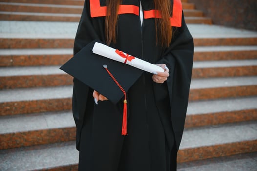 Happy cute brunette caucasian grad girl is smiling. She is in a black mortar board, with red tassel, in gown, with nice brown curly hair, diploma in hand. photo