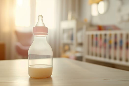Baby transparent feeding bottle with milk on the table in a bright room, in the background there is an out of focus baby crib photo