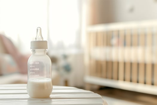 Baby transparent feeding bottle with milk on the table in a bright room, in the background there is an out of focus baby crib photo