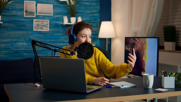 Influencer with headphones using phone for taking selfie recording podcast series for audience. On-air online production internet broadcast show host streaming live content for digital social media photo