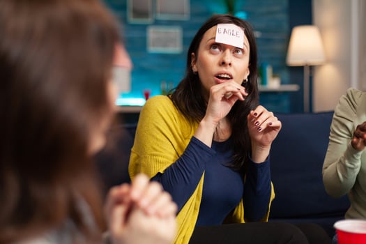 Mixed race friends with sticky notes attached on forehead playing guess who game during home party. Multi-ethnic people having fun, laughing together while sitting on sofa in living room late at night. photo
