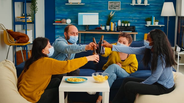 Mixed race friends clinking beer bottles hanging out keeping social distance having fun in living room during global pandemic. Diverse group of people celebrating with toast in outbreak enjoying time photo