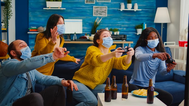 Excited multiethnic friends trying to win games enjoying new normal party during global pandemic wearing face mask, keeping distancing sitting on couch in living room supporting women photo