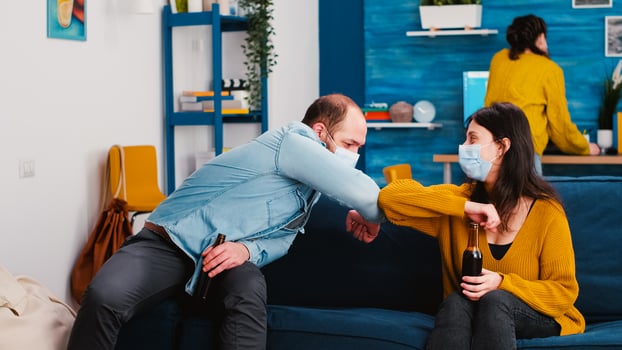 Joyfull friends sitting on couch in living room during new normal party making new greeting elbow bump avoiding spread of coronavirus. People talking, enjoying a beer, spending time together photo