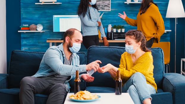 Mixed races friends with medical masks disinfecting hands with sanitizer gel spending time together in living room. Diverse people talking during party, enjoying beer and snacks in global pandemic photo