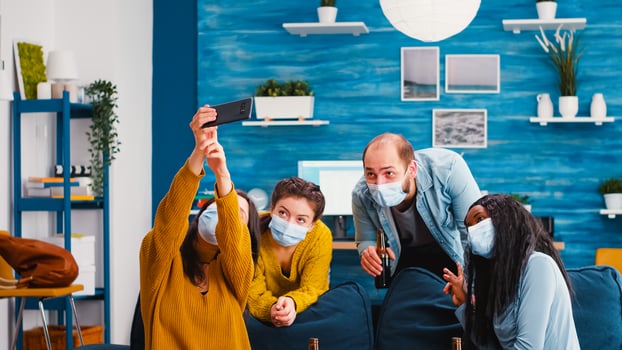 Young people from diverse cultures taking selfie wearing protection masks having fun together during new normal party keeping social distancing, holding bottles of beer enjoying time in living room photo