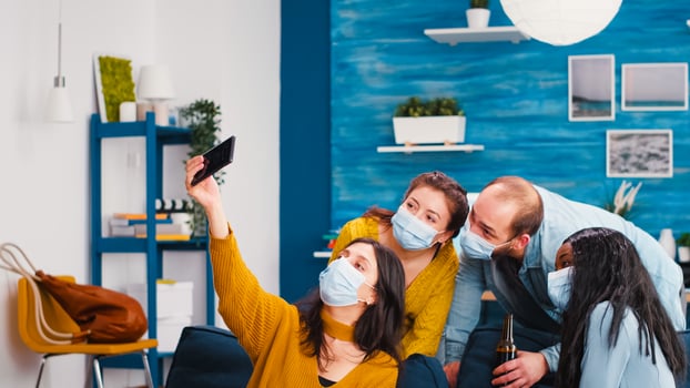 Multi-ethnnic friends with protective masks partying with beer taking selfie during covid 19 outbreak, new normal lifestyle concept with people having fun in living room respecting social distance photo
