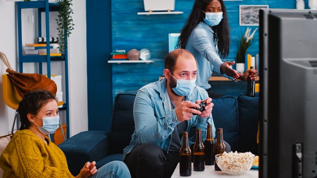 Group of mixed races friends playing game in home living room with joystick wearing face mask keeping social distancing in time of corona outbreak. Diverse people enoying beer and chips. photo