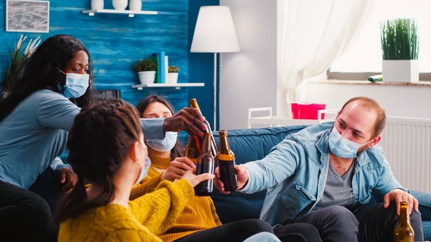 Young people toasting and cheering bottles of beer spending free time in living room respecting social distance to prevent virus spread. Diverse friends enjoying new normal party in outbreak photo