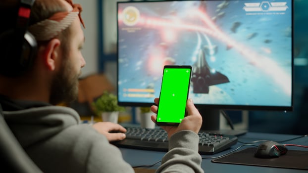 Concentrate gamer holding phone with green screen mock up chorma key display while playing games on powerful computer streaming online competition. Player using isolated desktop playing shooter games photo