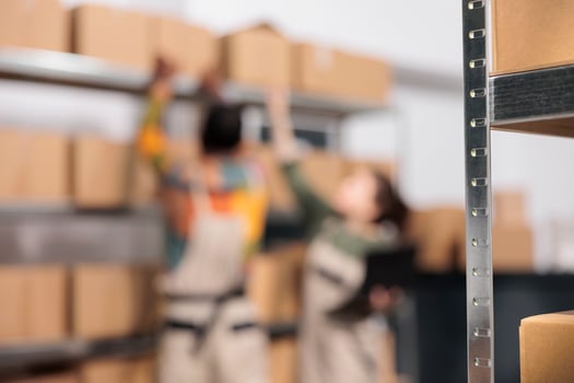 Selective focus of carton boxes standing on shelves, in background storehouse employees preparing customers orders. Storage room team managing parcels transportation, working in warehouse photo