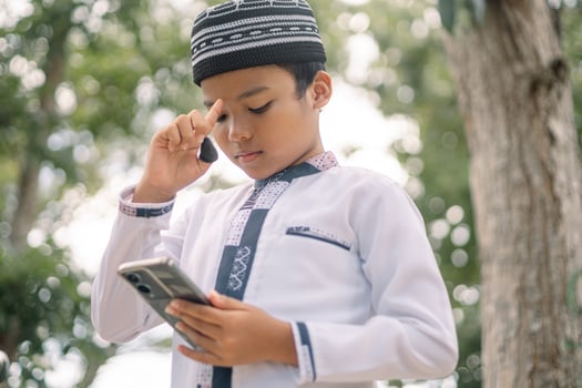Baubau, Indonesia, June 15 2024 - Asian Muslim boy wearing a cap is playing with his smartphone photo