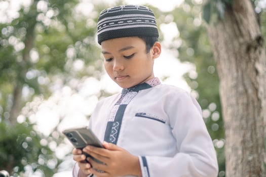 Baubau, Indonesia, June 15 2024 - Asian Muslim boy wearing a cap is playing with his smartphone photo