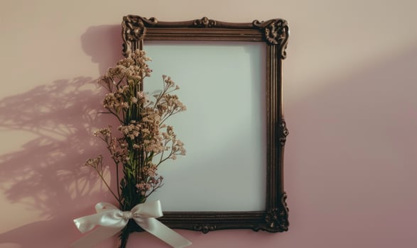 White blank card in a vintage bronze frame on a pastel lilac background, with a small bunch of wildflowers and a ribbon photo
