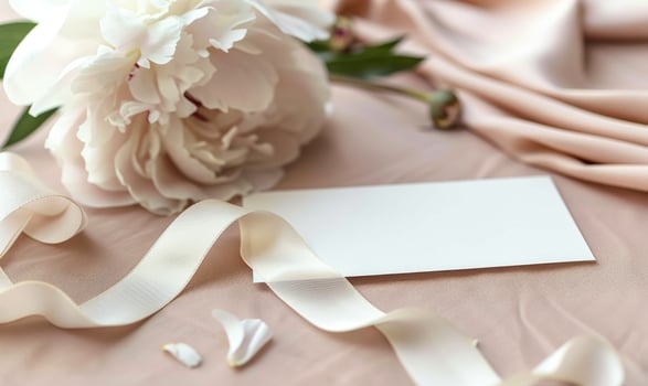 White blank card on a pastel rose pink table, with a piece of satin ribbon and a single peony flower photo