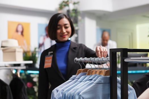 Clothing store smiling asian woman seller counting shirts hanging on rack while managing apparel inventory. Department center boutique worker checking merchandise on hangers photo