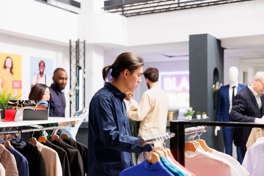Pensive Asian millennial guy customer shopping in apparel store. Confused young man choosing and buying new clothes in shopping mall, looking through male clothing items on hangers photo