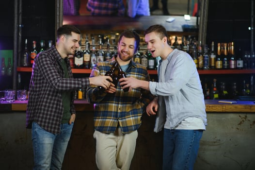 Three young men in casual clothes are smiling and clanging glasses of beer together while sitting at bar counter in pub photo