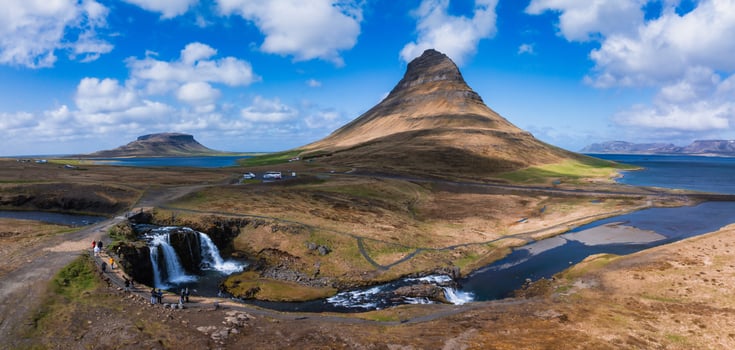 Stunning aerial panorama view of Kirkjufell mountain in Iceland, showcasing its distinctive shape. The image features a parking lot with cars, blue sky, and deep blue water. photo