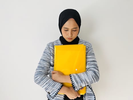 An upset young Asian woman holds a file folder in front of her chest. Business woman sad because of failed attempt. Female wear black headscarves and blazers photo