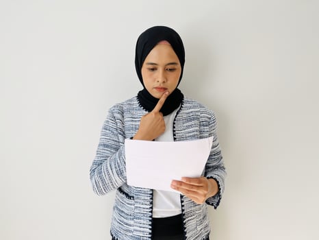 Young Asian woman thinking while reading document. Standing read report, planning focusing on contract. Female professional wearing black hijab and tweed blazer isolated by white background. photo