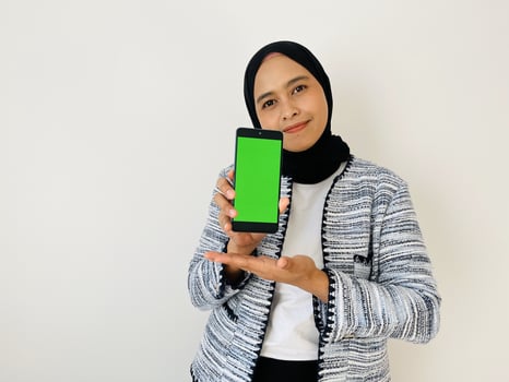 A smiling young Asian Muslim woman show smartphone green color of the screen with positive expression, dressed in tweed blazer and black hijab feeling happy isolated by white background. Mobile phone photo